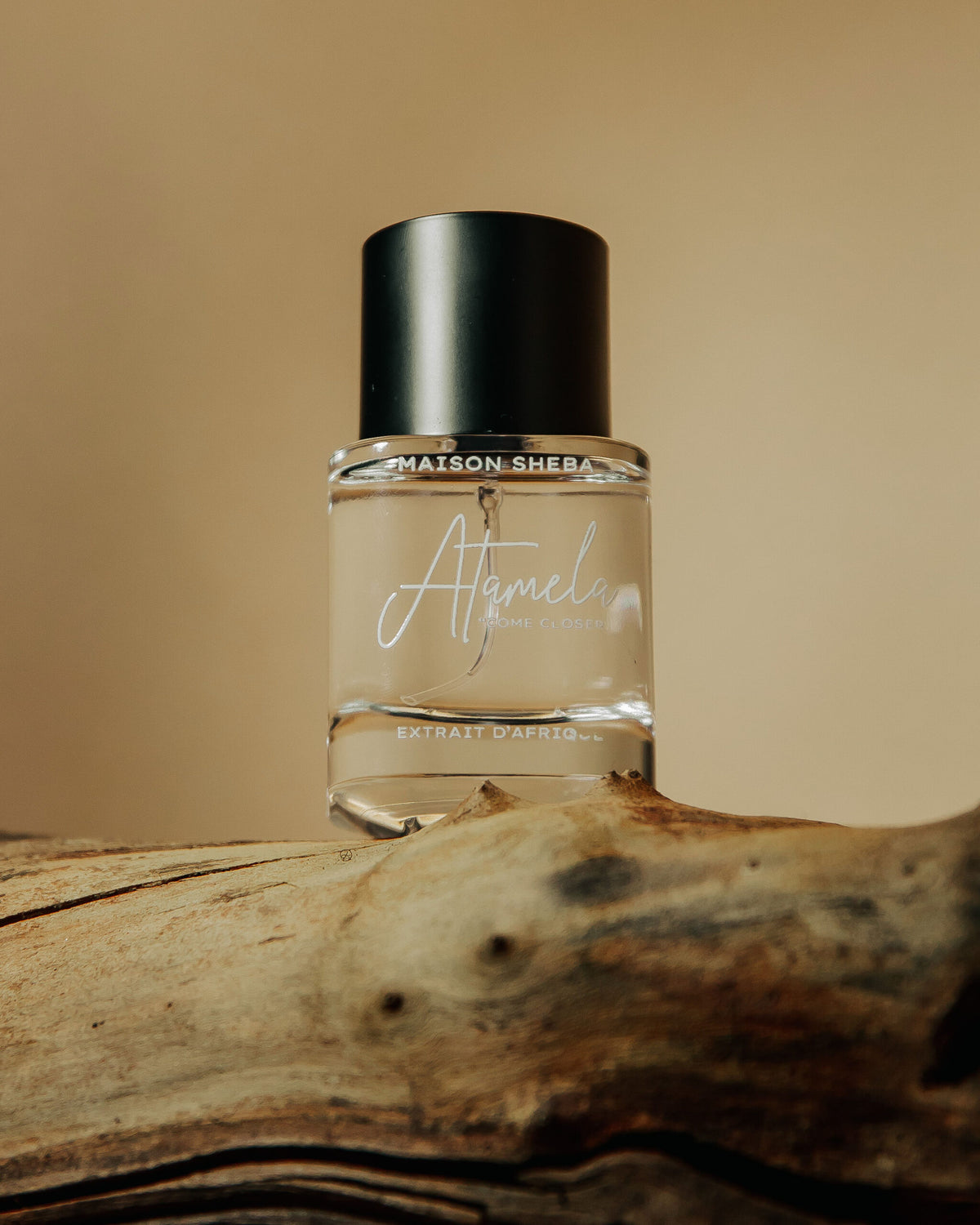 Maison Sheba Atamela Extrait d’Afrique, a clear glass bottle with a black cap and a script Atamela label subtitled Come Closer, resting on driftwood