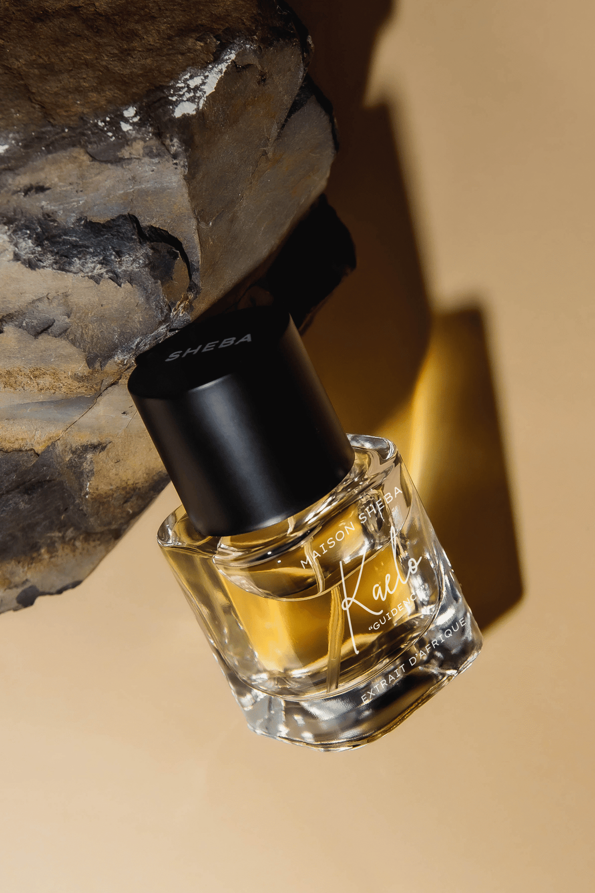 Maison Sheba Kaelo Extrait d’Afrique, a tilted glass bottle of golden liquid with a black Sheba cap, leaning against grey rock in warm light