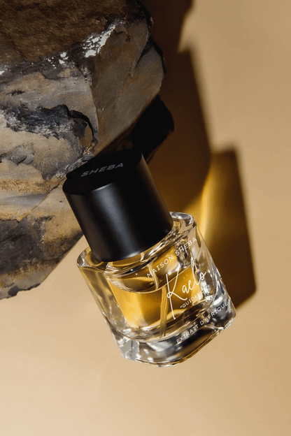 Maison Sheba Kaelo Extrait d’Afrique, a tilted glass bottle of golden liquid with a black Sheba cap, leaning against grey rock in warm light